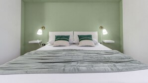 Un letto rifatto con cura, una coperta grigia, due cuscini verdi e due abat-jour su comodini bianchi.