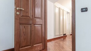 Una porta in legno con una serratura, un corridoio con pavimento in legno e un termostato fissato al muro.