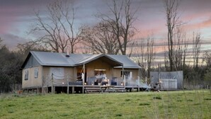 Un chalet rustique doté d’un porche et d’un espace extérieur aménagé pour s’asseoir, entouré d’arbres et d’un champ verdoyant.