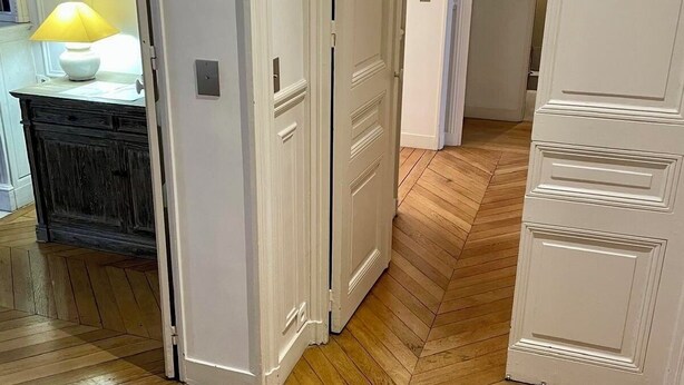 Ein Zimmer mit Fischgrätparkett, weißen Türen und einer Lampe auf einem dunklen Holzschrank.