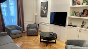 Ein modernes Wohnzimmer mit Fischgrätparkett, grauen Sofas, einem schwarzen Couchtisch, einem an der Wand montierten Fernseher und einem Bücherregal mit Büchern und Dekoration.
