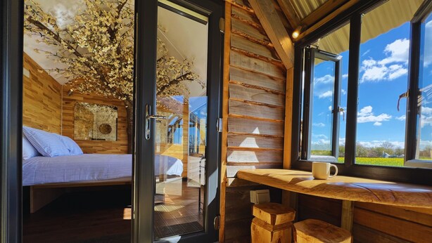 Ein Holzblockhaus-Interieur mit einem Bett, einem Tisch mit Hocker, einer Lampe und Blick ins Freie.