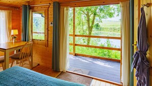 Een houten kamer met een bed, een bureau en een stoel. Er is een raam met gordijnen en uitzicht op begroeiing.
