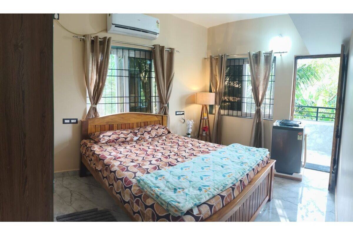 A bedroom with a bed, curtains, an air conditioner, a lamp, and a view of greenery outside.