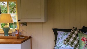 Ein Zimmer mit Holztisch, einer Lampe, einem Fenster mit Blick auf Grünflächen, einem Bett mit floralem Bettzeug und einem karierten Kissen.