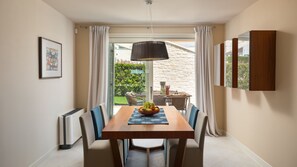A dining area with a wooden table, chairs, a fruit bowl, and a view of a garden through a glass door.