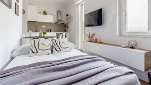 Ein modernes Schlafzimmer mit einem großen Bett, einem Nachttisch, einem an der Wand montierten Fernseher und Blick auf die Küche im Hintergrund.