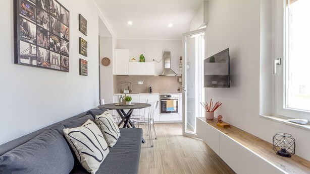 Eine moderne Wohnung mit einem grauen Sofa, einem Esstisch, einer Küche mit weißen Schränken und einem an der Wand befestigten Fernseher.
