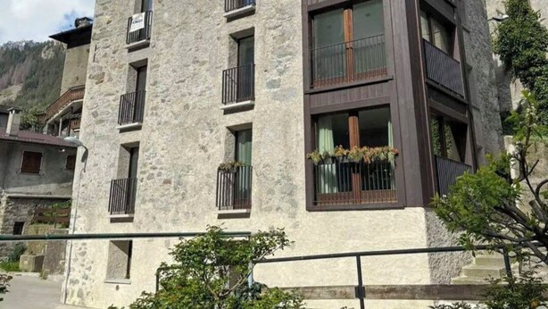 Ένα πολυώροφο κτίριο με πέτρινη πρόσοψη και μπαλκόνια, το οποίο περιβάλλεται από πλούσια βλάστηση και βουνά.