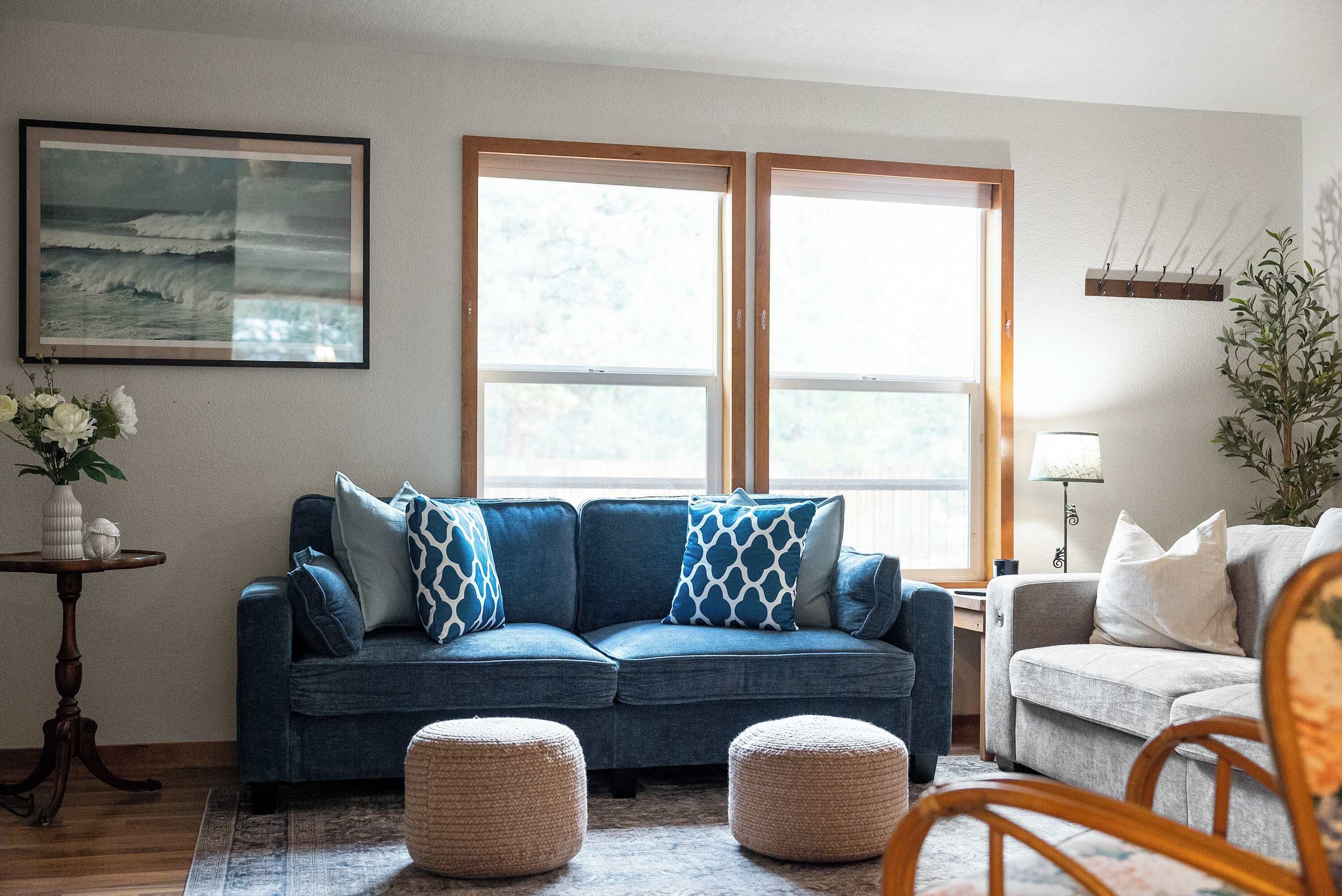 Un salon avec un canapé bleu, deux poufs, une table basse, un tableau encadré au mur et une fenêtre donnant sur des arbres.