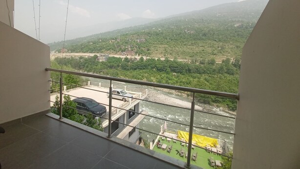 A balcony with a view of a river, greenery, and distant buildings. - Diamond Vaishali (Kullu)