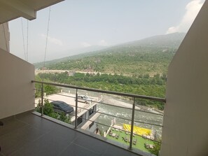 A balcony with a view of a river, greenery, and distant buildings. - Diamond Vaishali (Kullu)