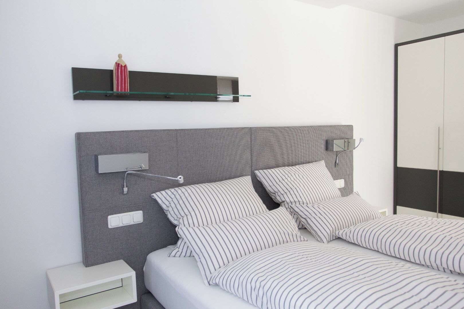 A modern bedroom with a grey headboard, a bedside table, a nightstand, a bedside lamp, and a wardrobe.