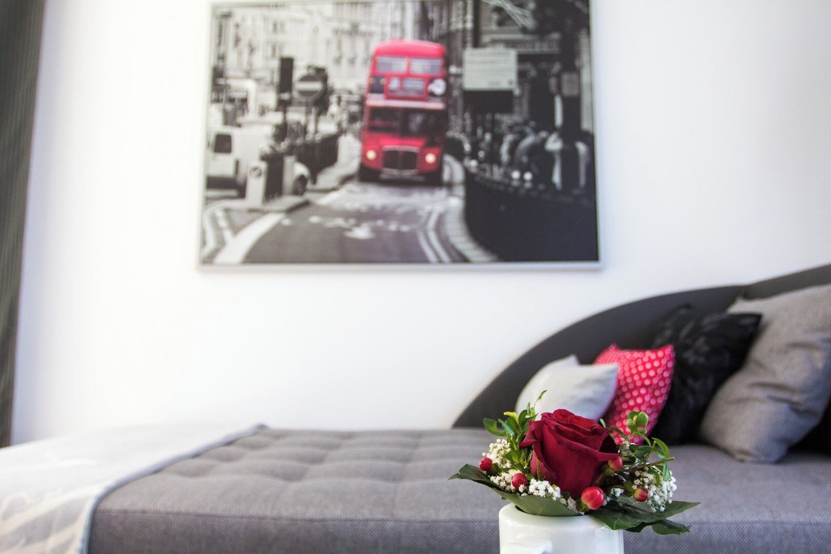 Un dormitorio con un sofá gris, un jarrón blanco con una rosa roja, y una fotografía blanco y negro de un autobús rojo de dos pisos en la pared.