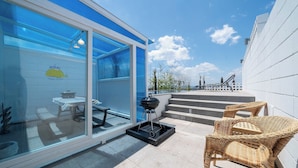 Eine moderne Terrassenanlage mit Glasveranda, Grill, Korbmöbeln und Blick auf das Schwimmbecken.