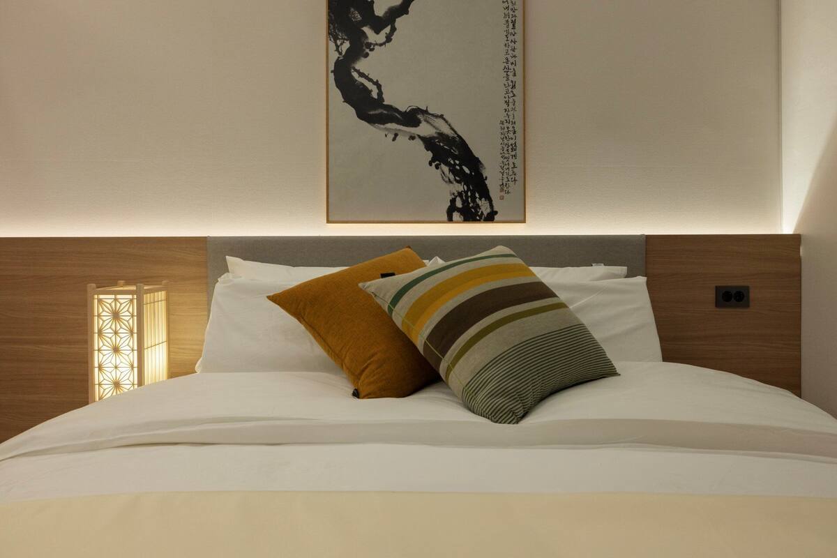 Un letto rifatto con lenzuola bianche e due cuscini decorativi, un’opera d’arte appesa al muro e una testiera in legno.