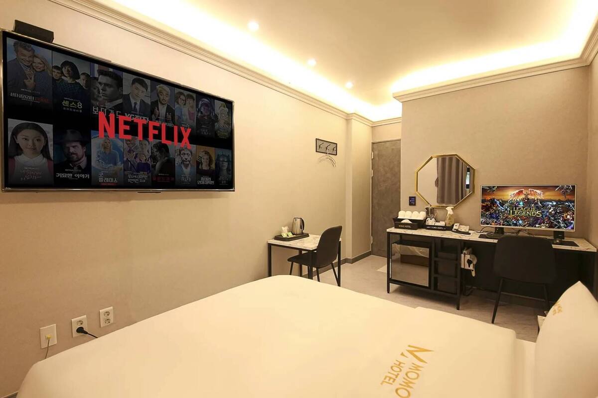 Una camera d'albergo con un letto grande, una scrivania con un computer, una TV a muro che mostra Netflix e uno specchio sopra la scrivania.