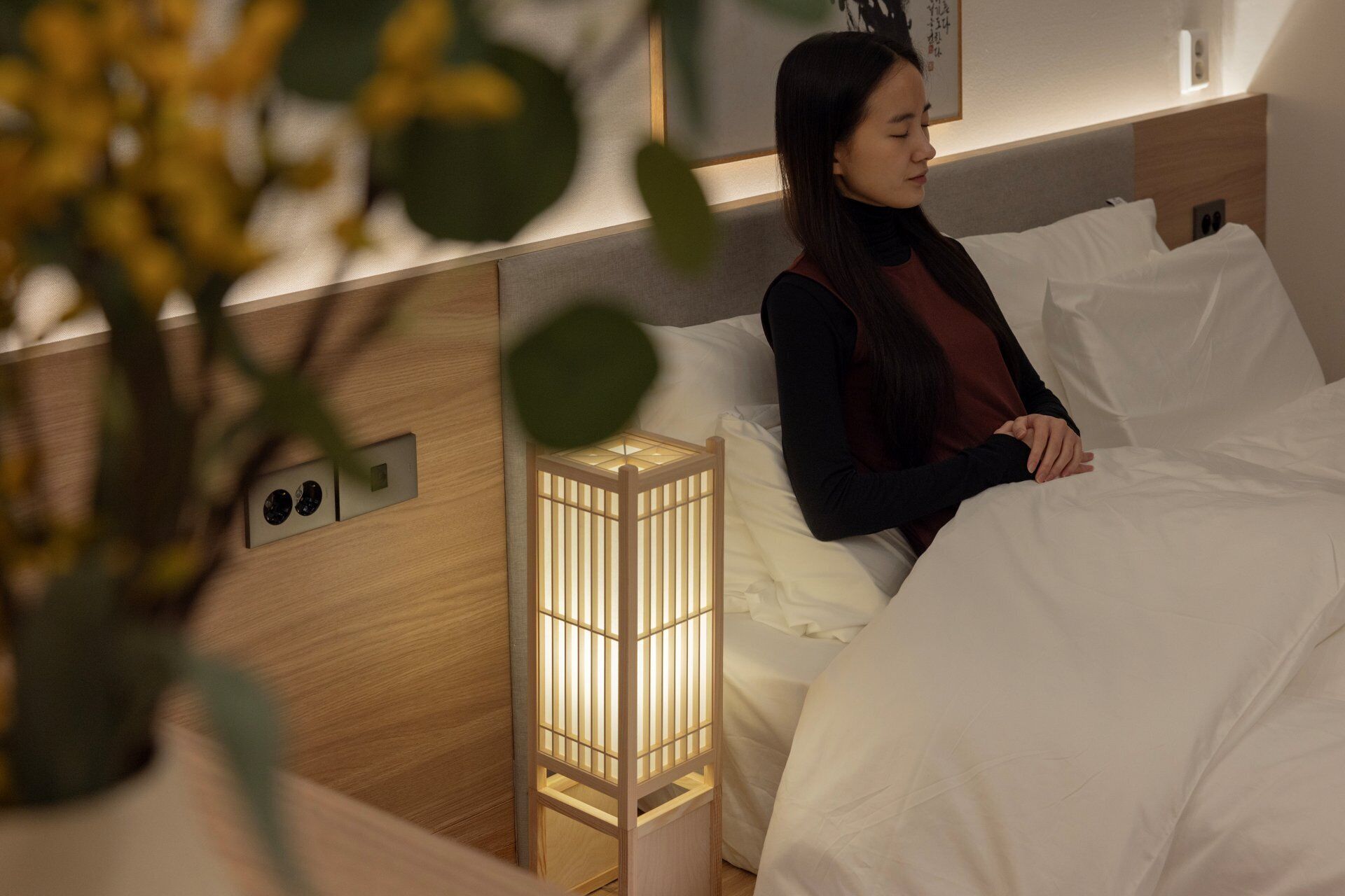 A hotel room with a bed, a bedside table, a wall-mounted light, and a power outlet.