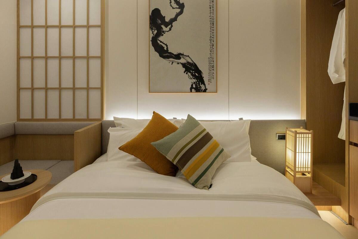Una camera d'albergo ordinata con un letto, cuscini, un comodino, un'opera d'arte appesa al muro e una tradizionale lanterna giapponese.
