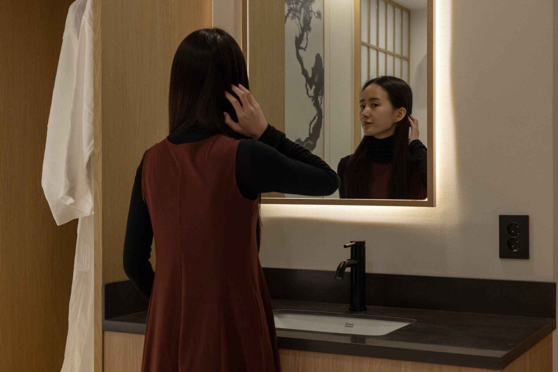 A person in a red dress standing in front of a bathroom mirror with a sink and a black faucet.