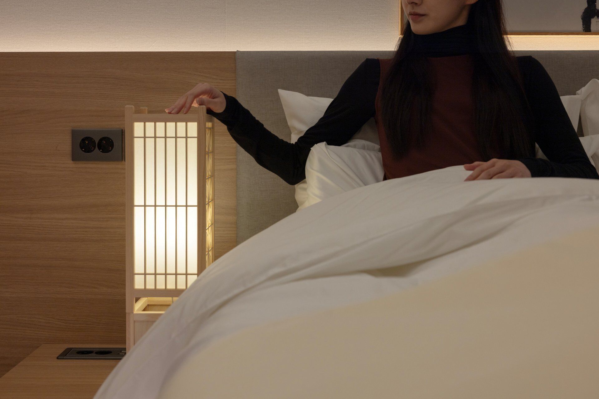Una persona en una habitación con una cama, una mesita de noche y una lámpara de pared.