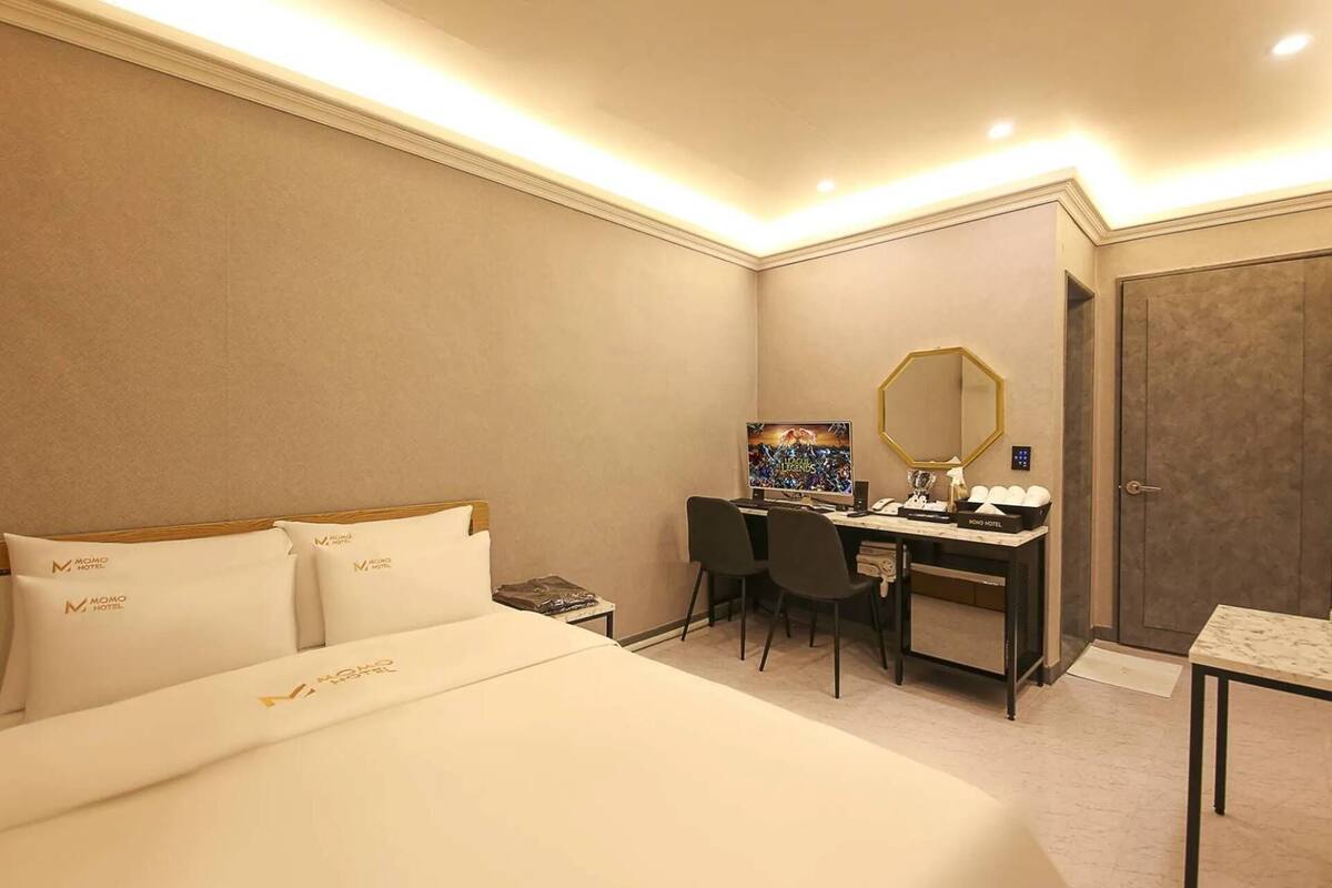Una camera d'albergo con un letto, una scrivania con un computer, una sedia, uno specchio e una porta.