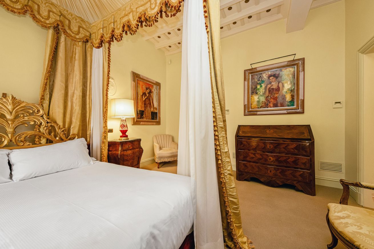 A bedroom with a four-poster bed, a wooden dresser, a chair, and two paintings on the walls.