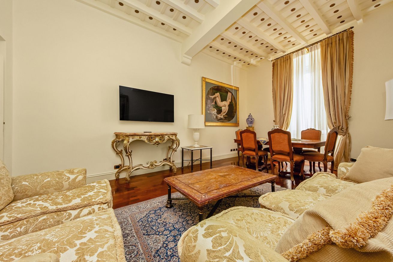 A living room with a flat-screen TV, a painting, a dining table with chairs, and patterned sofas.