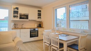 A modern kitchen with white cabinetry, a dining table with chairs, and a large window with a view of buildings. - Südstrand Apartment - your modern hideaway for a relaxing Baltic Sea vacation (Grömitz)