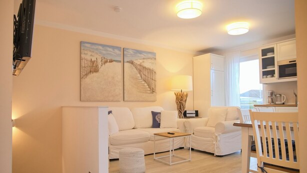A modern living room with a sofa, armchairs, a coffee table, and a dining area with a chair. There are two paintings on the wall and a lamp on a side table. - Südstrand Apartment - your modern hideaway for a relaxing Baltic Sea vacation (Grömitz)