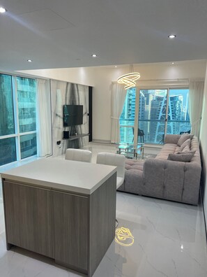 A modern living room with a large sofa, a dining area with a glass table, and a TV mounted on the wall. - Stunning Modern 1BR Apartment  Lake View JLT – Lake Terrace (دبي)
