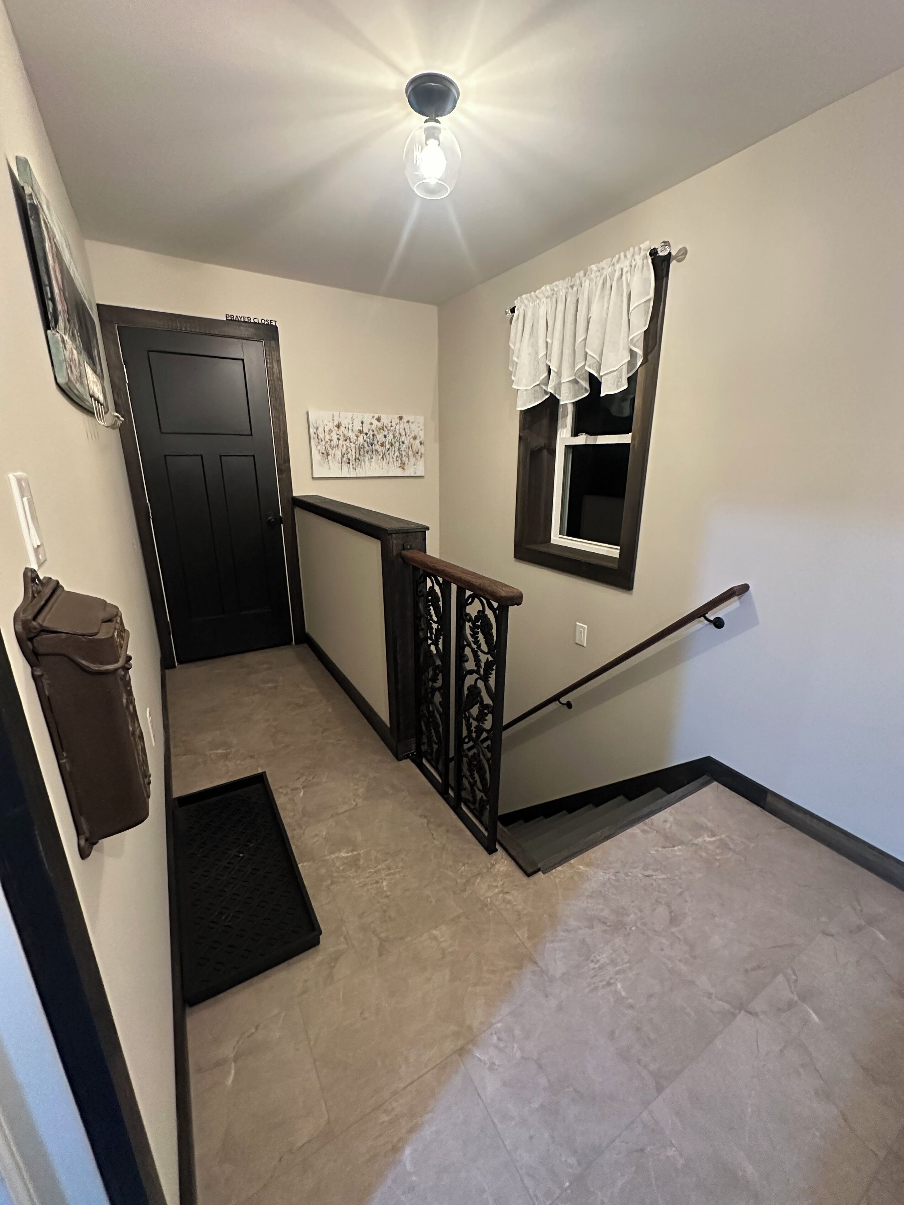 A hallway with a door labeled "PRAYER CLOSET," a window with white curtains, a wall-mounted artwork, and a staircase with a black railing.