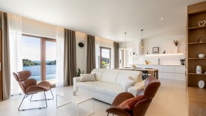 A modern living room with a white sofa, two brown armchairs, a glass coffee table, and a view of a lake through large windows. - Villa Cristal Blue by Villas Guide (Blato)