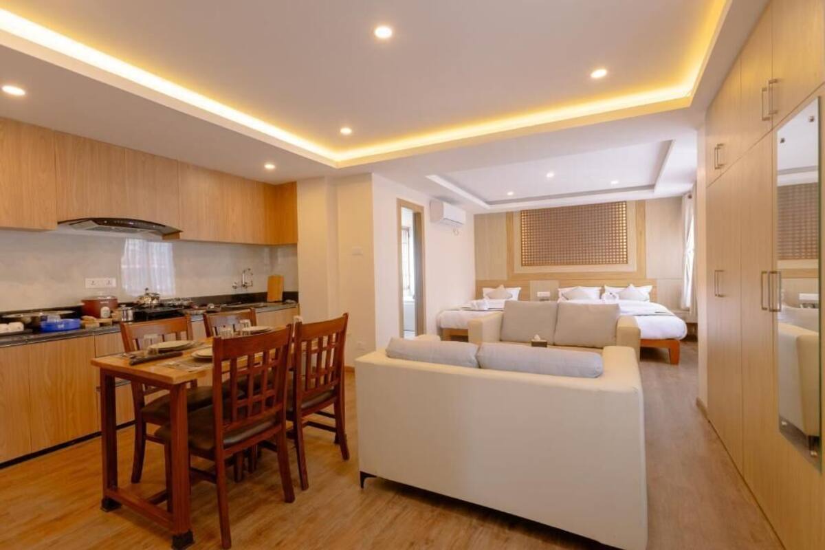 A modern kitchen with wooden cabinets, a dining table with chairs, and a white sofa in a living area with a bed and a window.