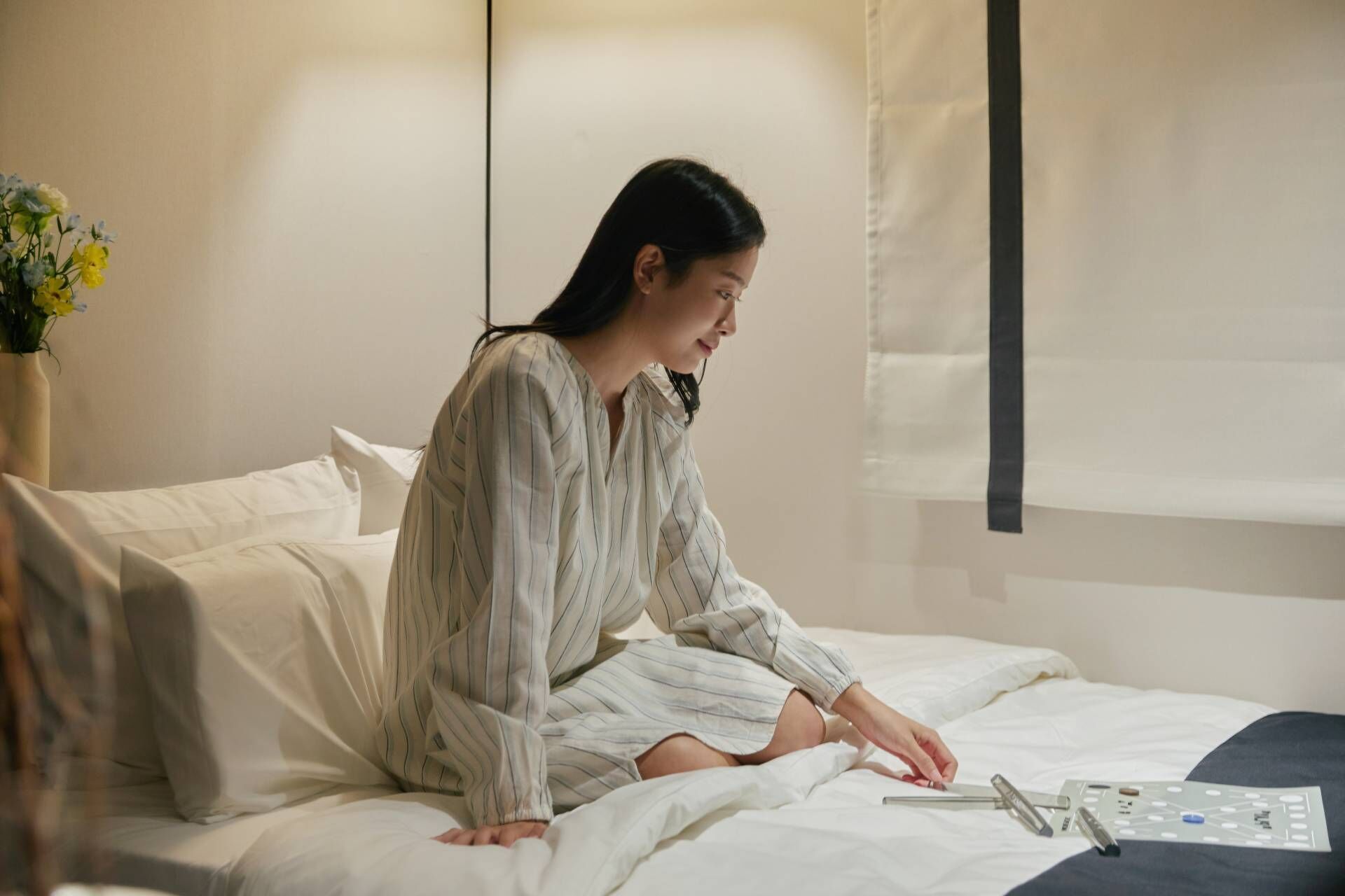 A person sitting on a bed in a room with a bedside table, a window with curtains, and a vase with flowers on a nightstand.