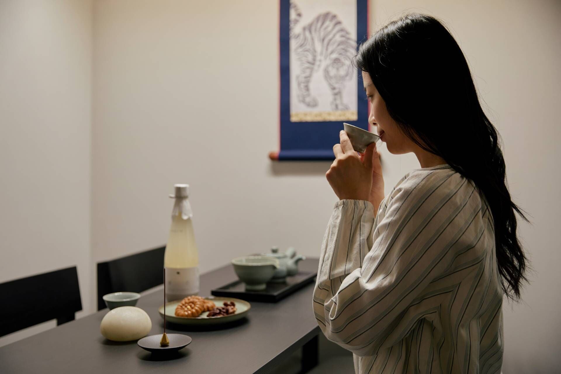 A person in a striped shirt standing at a table with a bottle, a teapot, and a plate of food.