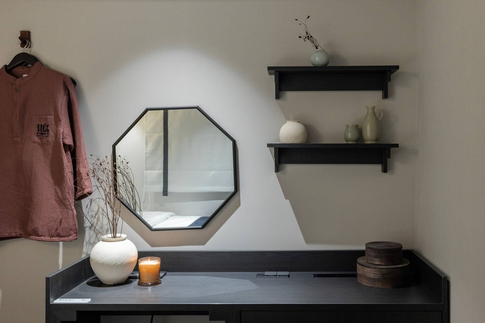 A modern room with a large octagonal mirror, a desk with a vase, candle, and a small plant. There are two black shelves above the mirror, each holding decorative items.