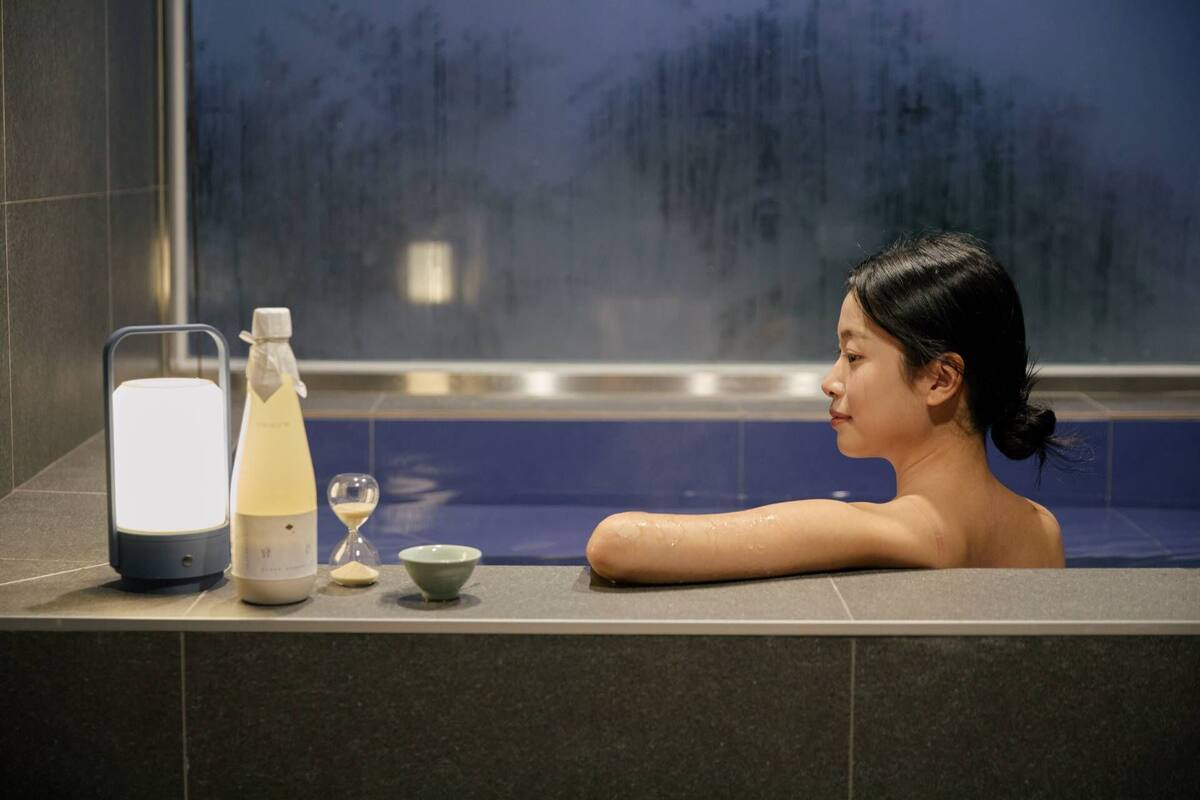 Eine Person in einer Badewanne mit einer Flasche, einem Glas und einem Behälter am Rand.