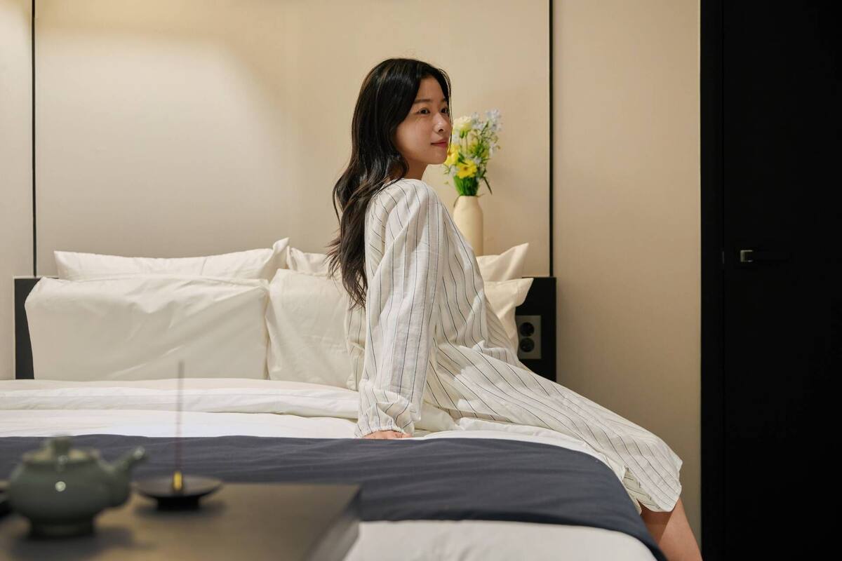 Eine Person sitzt auf einem Bett in einem Zimmer mit weiß-schwarzer Farbgestaltung, einer Vase mit Blumen und einem Teekessel auf dem Nachttisch.