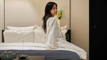 A person sitting on a bed in a room with a white and black color scheme, a vase with flowers, and a teapot on the bedside table.