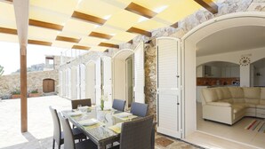 Ett takförsett utomhusområde med plats för måltider, med väggar av sten och utsikt över ett vardagsrum med en beige soffa.