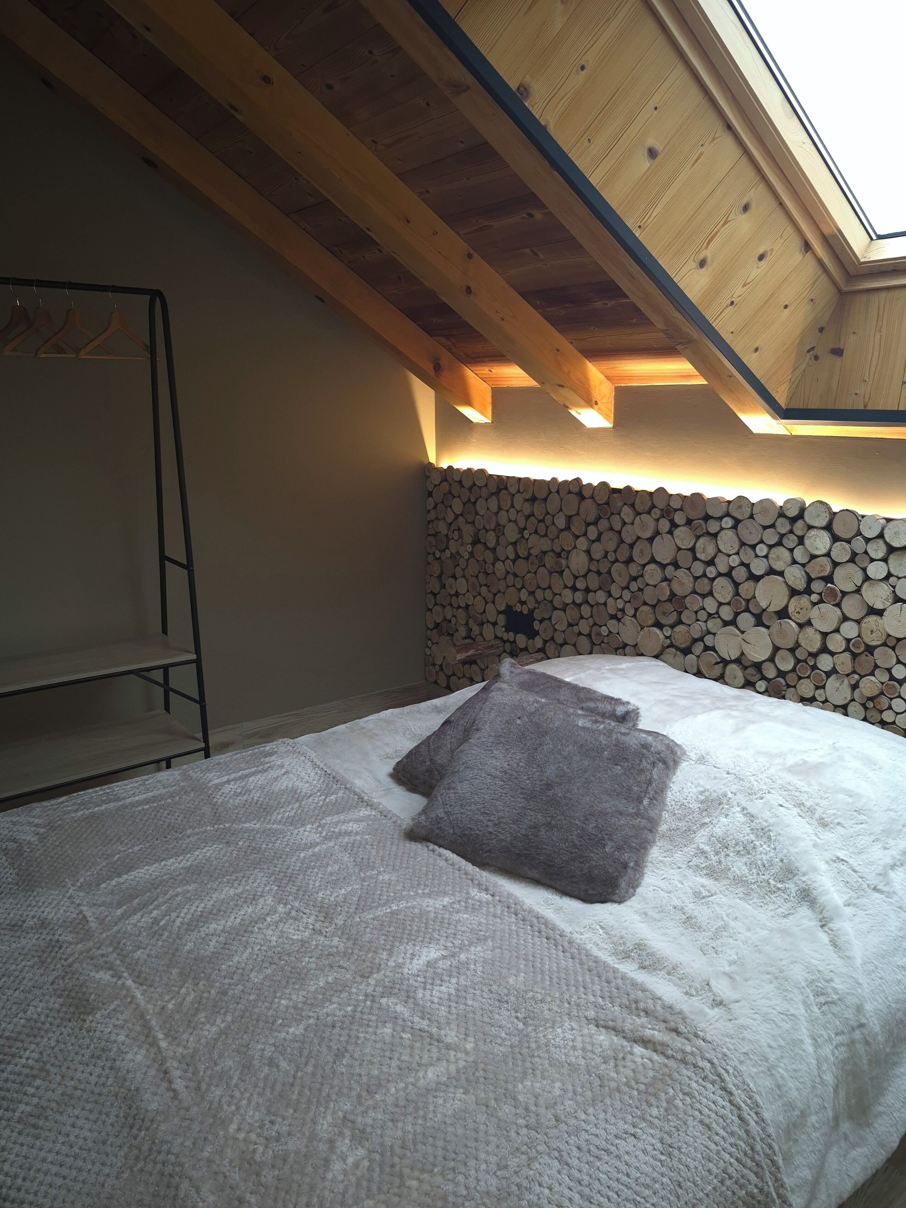 A bedroom with a sloped ceiling, a wooden beam structure, a bed with a white duvet and a gray pillow, and a wall of stacked logs.