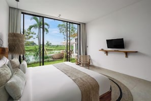 Une chambre avec un grand lit, un téléviseur posé sur une étagère et une vue sur un palmier et l’eau.