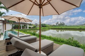 Un espace aménagé au bord de la piscine, avec des chaises longues et des parasols, une piscine, et une vue sur un plan d’eau et la végétation environnante.