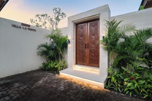 Entrada de una villa con un doble portón, camino de entrada pavimentado y exuberante vegetación.