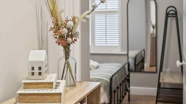 A bedroom with a bed, a bedside table with books and a vase of flowers, a window with blinds, and a full-length mirror. - 2BR Ground Floor • Parking • Central By Tempstay (Windsor)