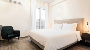 Hotellihuone, jossa on suuri sänky, tummanvihreä nojatuoli, yöpöytä lampun kanssa ja ikkuna, jossa on verhot.