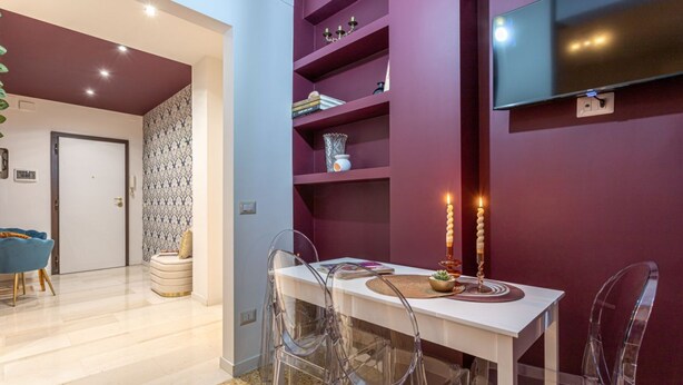 Un espace repas moderne avec une table blanche et des chaises transparentes, un mur d’accent violet et un téléviseur à écran plat fixé au-dessus de la table.