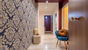 Un couloir avec un papier peint à motifs, une chaise bleue avec un coussin marron et une porte dotée d’une poignée en laiton.