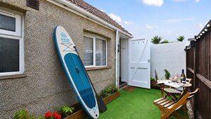 A house with a surfboard leaning against the wall, a small patio with a table and chairs, and a garden with plants. - Stone's Throw - a holiday let that sleeps 2 guests in 1 bedroom (Hythe)